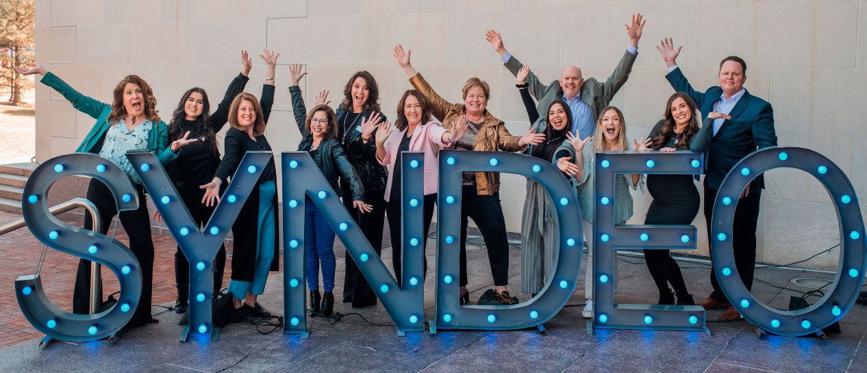 Syndeo Institute staff cheering the launch standing behind a lighted Syndeo marquee