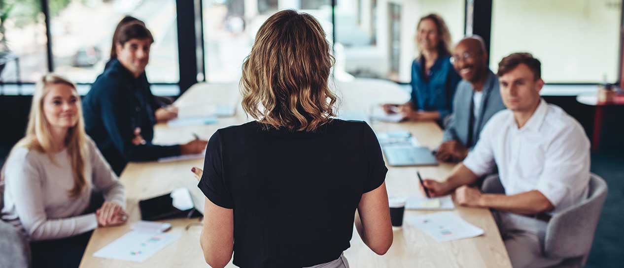 Five people at a conference table listening to a young woman standing at the end of the table with her back to us.