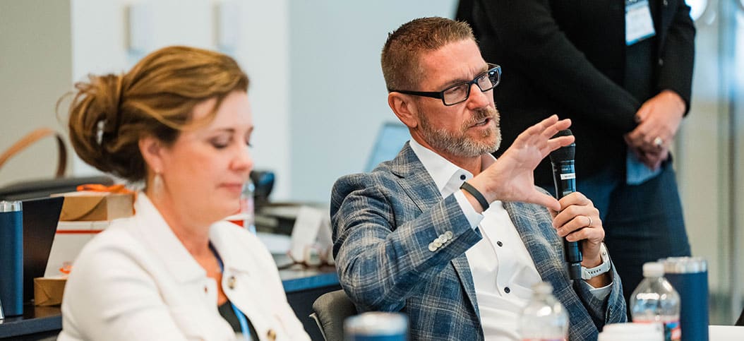 At the CXC Conference, a man in a suit speaks into a microphone while gesturing with his hand. A woman sits next to him, listening intently.