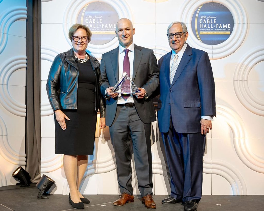 Three people in formal attire stand together as Jake Perlman in the center holds a triangular Innovation & Intrapreneurship Award for Excellence; a "Cable Hall of Fame" sign is visible in the background.