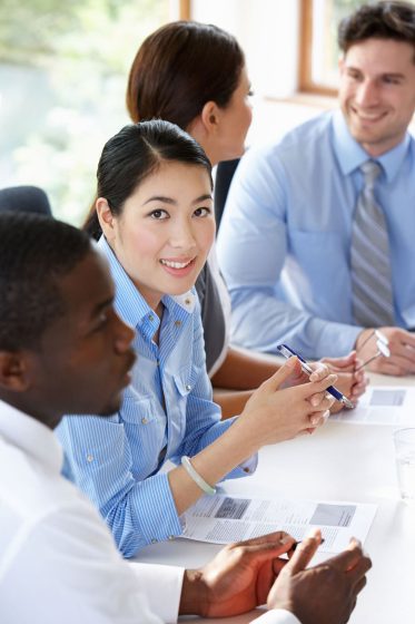Four people sit at a table in a meeting, holding pens and documents. One woman in the foreground looks at the camera while the others are engaged in conversation.